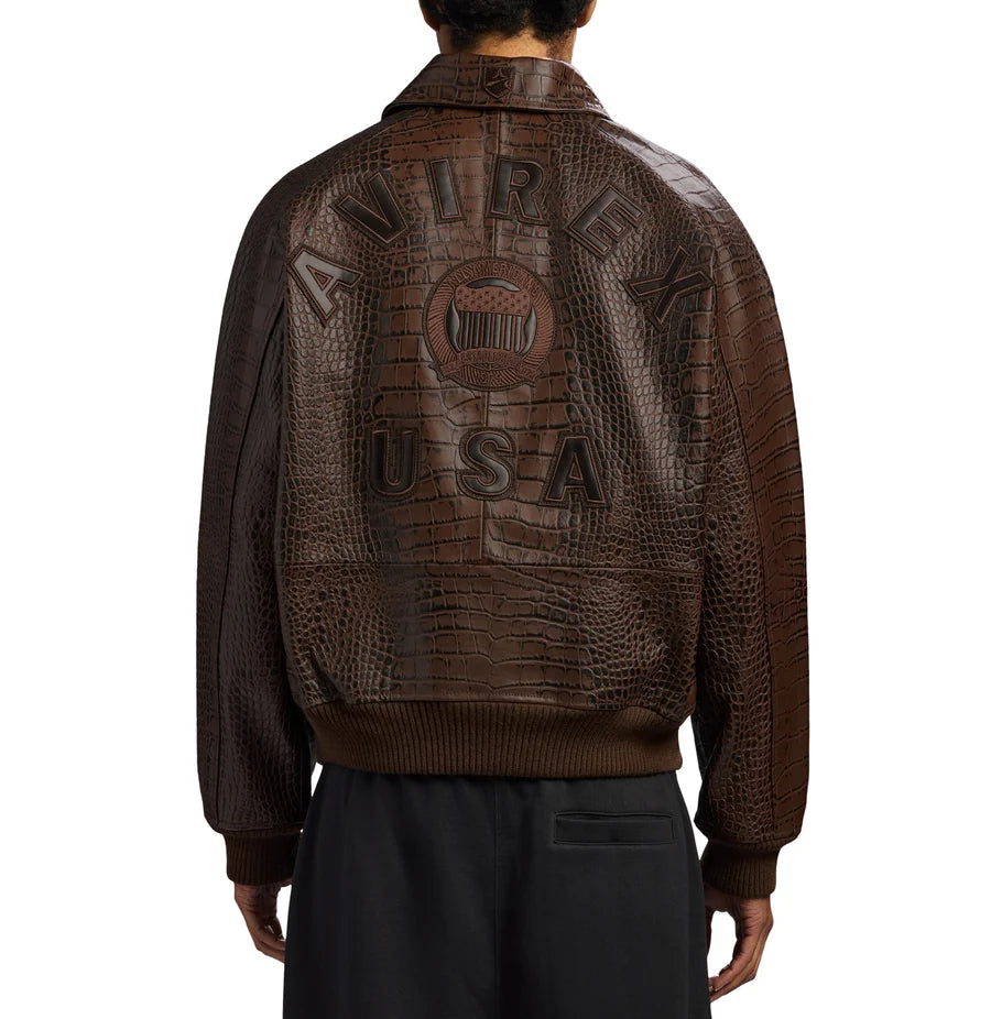 Back view of a men's brown crocodile-embossed AVIREX-style jacket featuring bold USA lettering, a vintage emblem, ribbed cuffs and hem, styled with black trousers.
