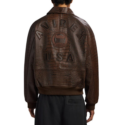 Back view of a men's brown crocodile-embossed AVIREX-style jacket featuring bold USA lettering, a vintage emblem, ribbed cuffs and hem, styled with black trousers.