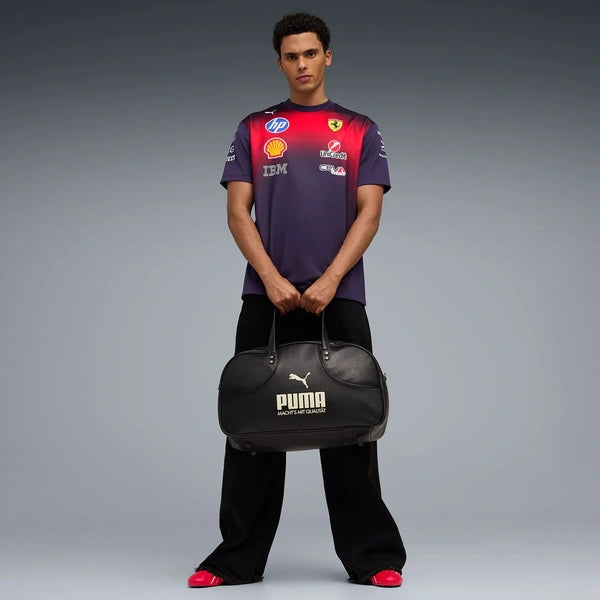 A man stands against a gray studio background wearing a purple and red Ferrari Formula 1 team t-shirt, black pants, and red shoes, while holding a black Puma duffel bag with the Puma cat logo and 'PUMA FORMULA 1' text.