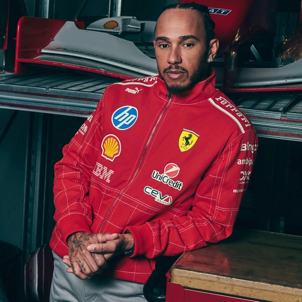 Lewis Hamilton wearing a red Ferrari team zip-up jacket with multiple sponsor logos, posed in a garage setting with a race car in the background.