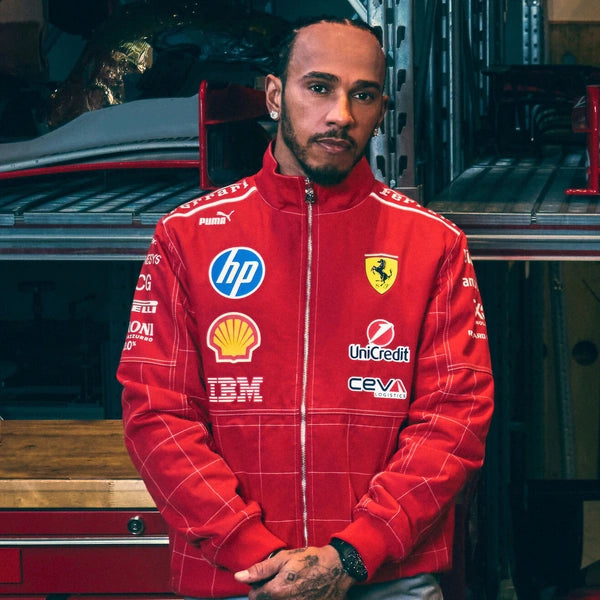 A man wearing a red Ferrari Scuderia Ferrari zip-up jacket with multiple sponsor logos, standing in front of a garage-like background with a car and equipment.