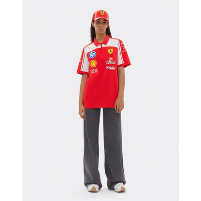 A model stands against a light gray studio background wearing a red and white Scuderia Ferrari short-sleeve polo shirt with multiple sponsor logos, a matching red Ferrari cap, loose dark gray pants, and white sneakers.