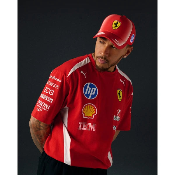 A man wearing a red Ferrari team cap and matching red sponsor jersey with white side panels and multiple team logos, posed against a dark studio background.