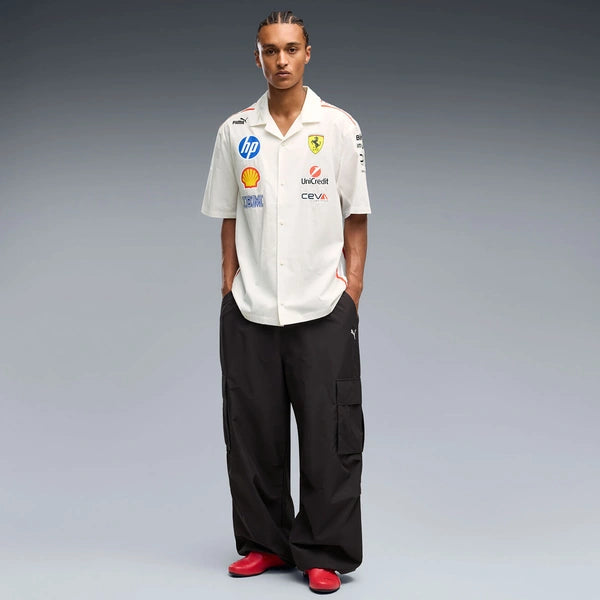 A man stands against a gray studio background wearing a white short-sleeve button-up shirt with multiple Ferrari and sponsor logos, black cargo pants, and red shoes.