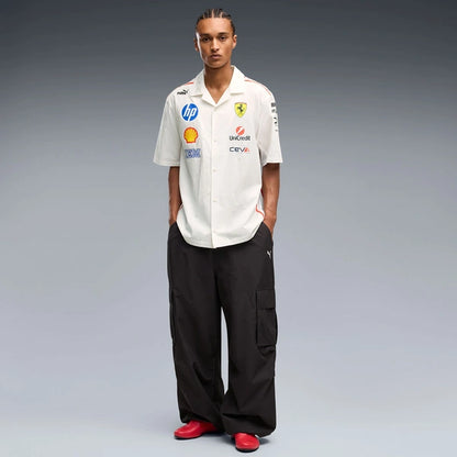 A man stands against a gray studio background wearing a white short-sleeve button-up shirt with multiple Ferrari and sponsor logos, black cargo pants, and red shoes.