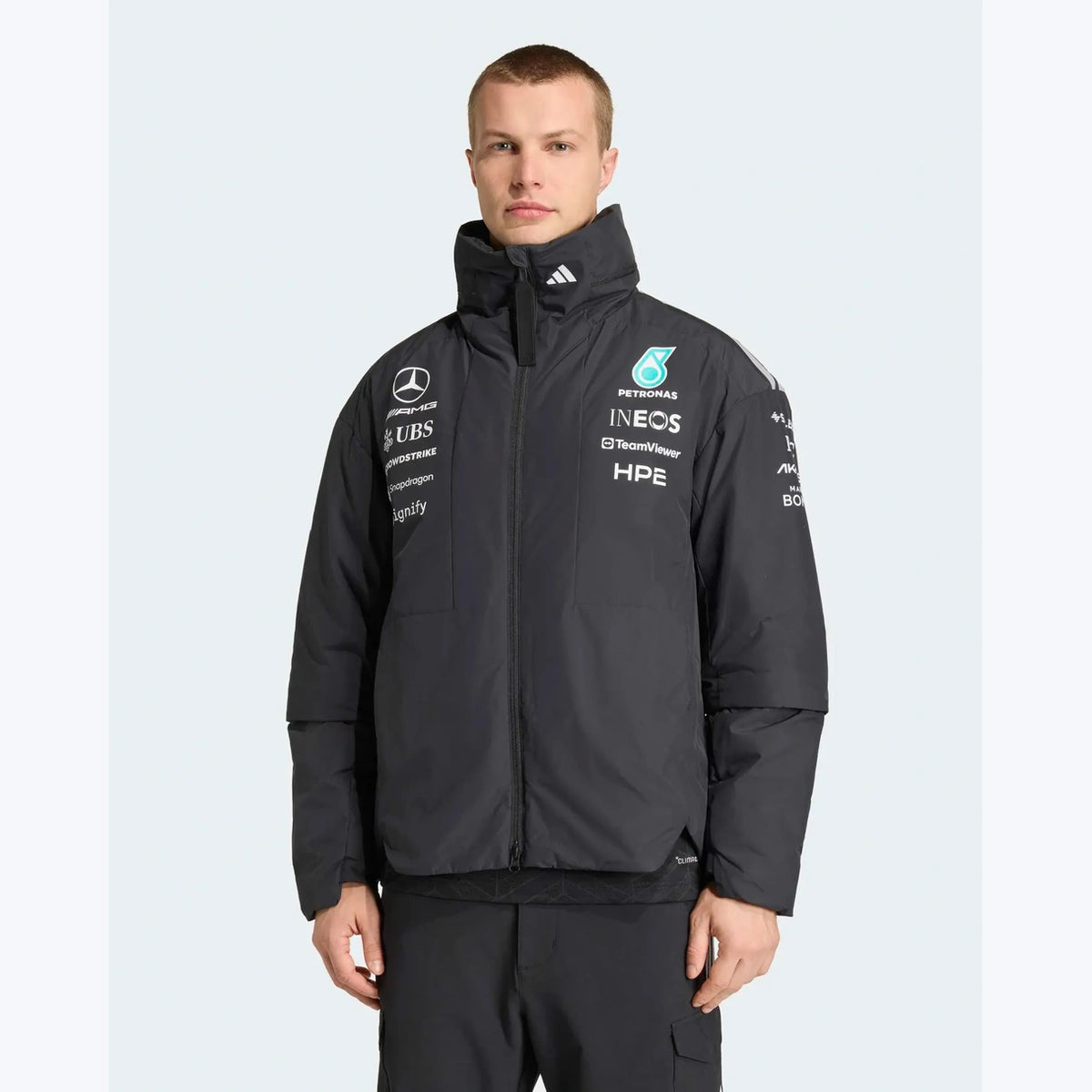 A man wearing a black Mercedes Formula 1 team puffer jacket with various sponsor logos on the chest, photographed against a light gray studio background.