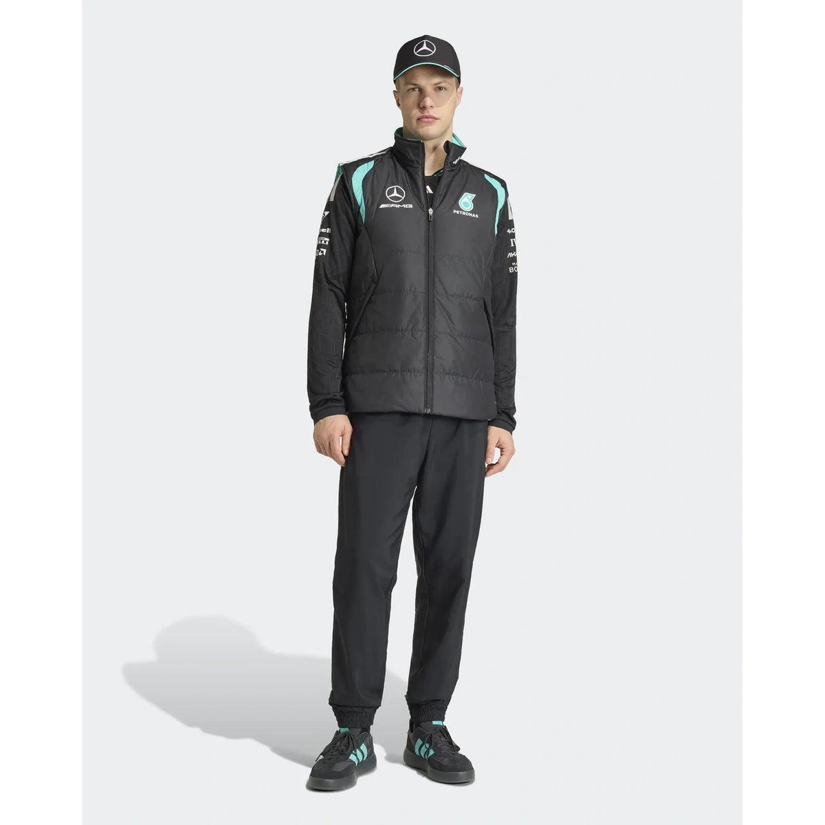 A male model wearing a black Mercedes Formula 1 team jacket with light blue accents, matching black trousers, a black cap, and black sneakers with light blue stripes, standing against a light gray studio background.