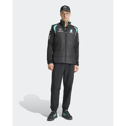 A male model wearing a black Mercedes Formula 1 team jacket with light blue accents, matching black trousers, a black cap, and black sneakers with light blue stripes, standing against a light gray studio background.
