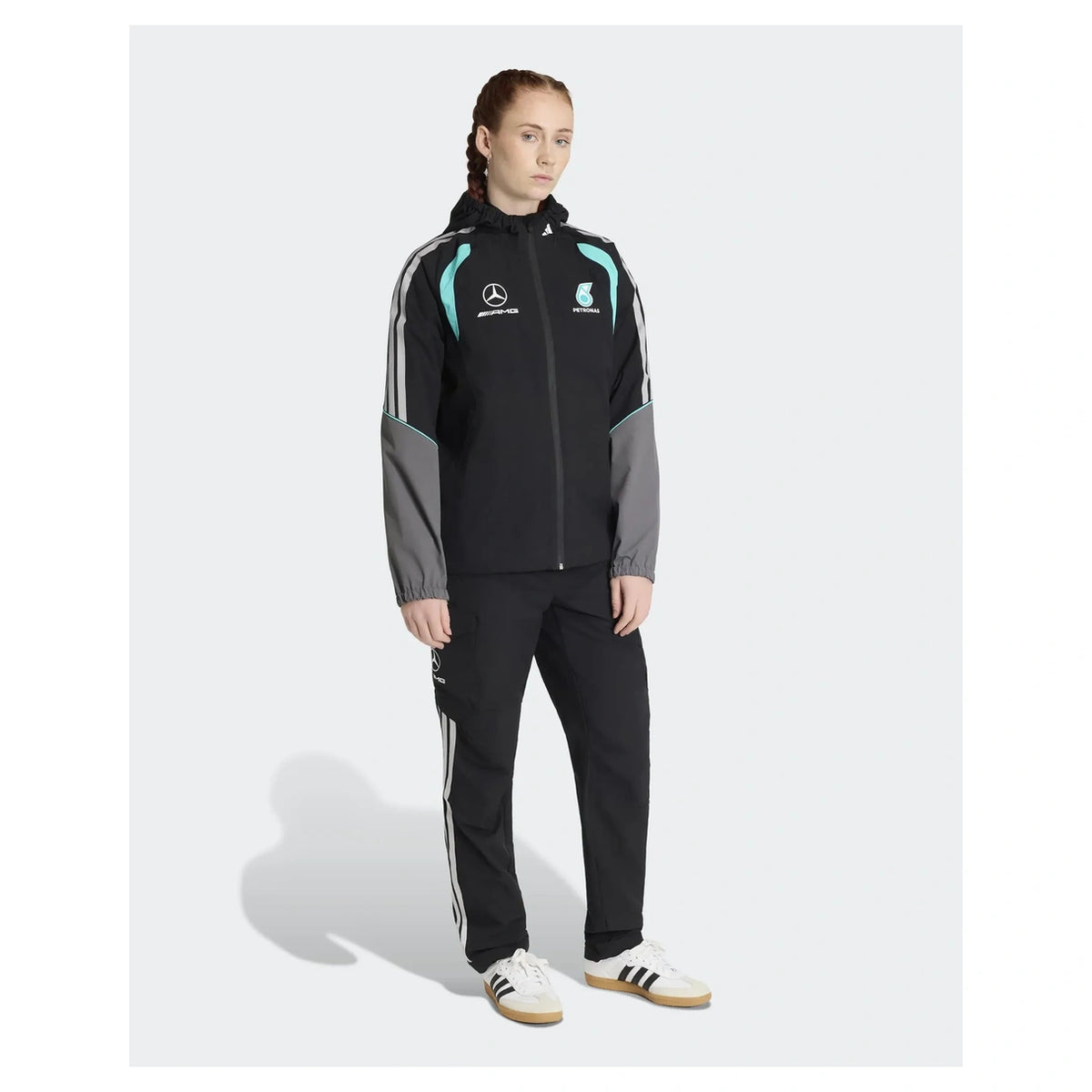 A model stands against a light gray studio background wearing a black Mercedes-AMG racing tracksuit with gray sleeves and teal accents, paired with black track pants and white Adidas sneakers.